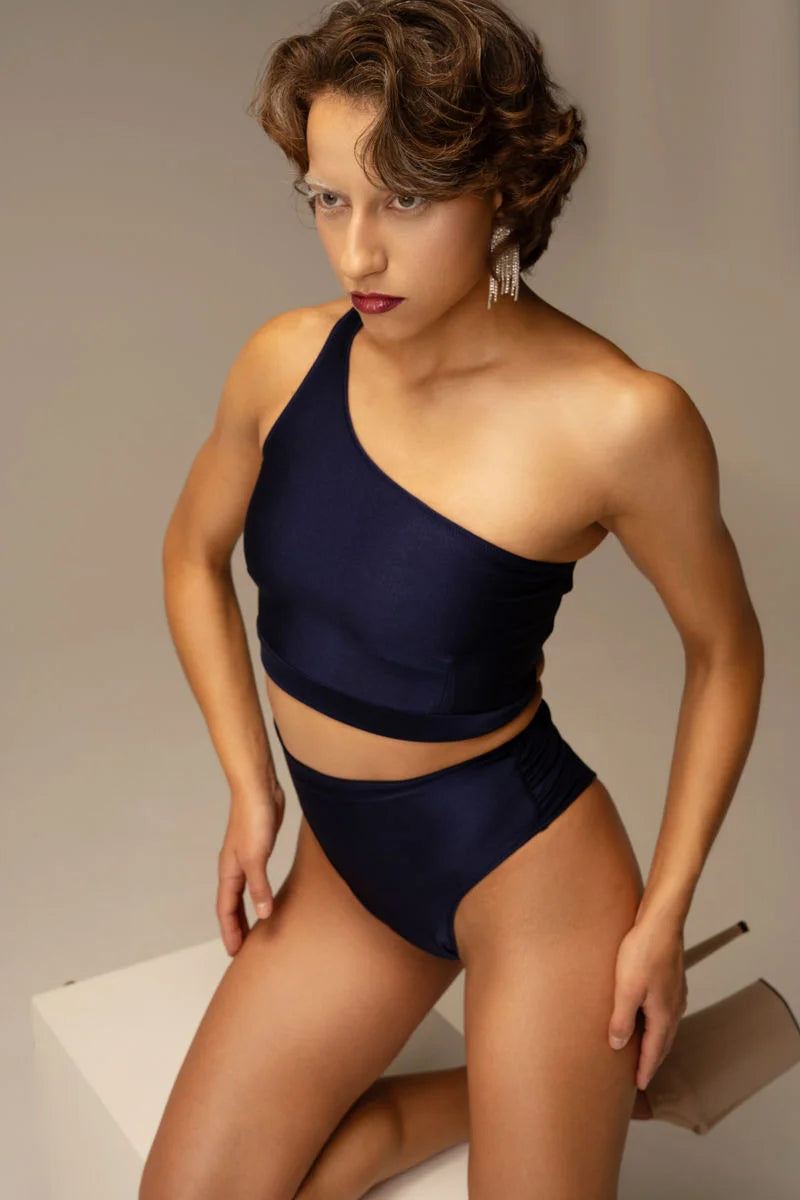 Model wearing Aura Top Sapphira and matching high-waisted shorts in shimmering sapphire blue, asymmetrical one-shoulder pole dance set, dynamic three-quarter view in studio.